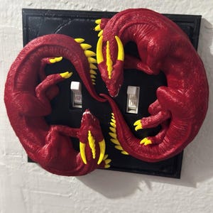 May include: A decorative light switch cover featuring two red dragons with yellow accents. The dragons are arranged in a circular pattern around the light switches, set against a black background. The switch plate is mounted on a white wall.