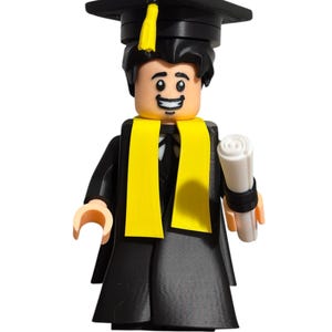 May include: A black graduation cap with a yellow tassel sits atop a smiling Lego figure wearing a black graduation gown and yellow scarf. The figure is holding a rolled-up diploma in its right hand.
