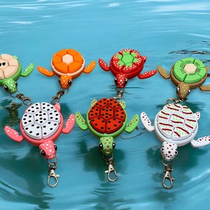 Puede incluir: Una colección de llaveros con forma de tortuga de colores. Cada llavero presenta un diseño diferente en su caparazón, incluyendo patrones que se asemejan a kiwi, sandía y otras frutas. Los llaveros tienen cierres metálicos y están sobre un fondo de agua azul.