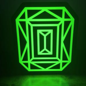 May include: A glowing green geometric design, resembling a gem, against a dark background. The neon-like light illuminates the intricate lines and shapes, creating a striking visual effect. The design is mounted on a dark, octagonal base.