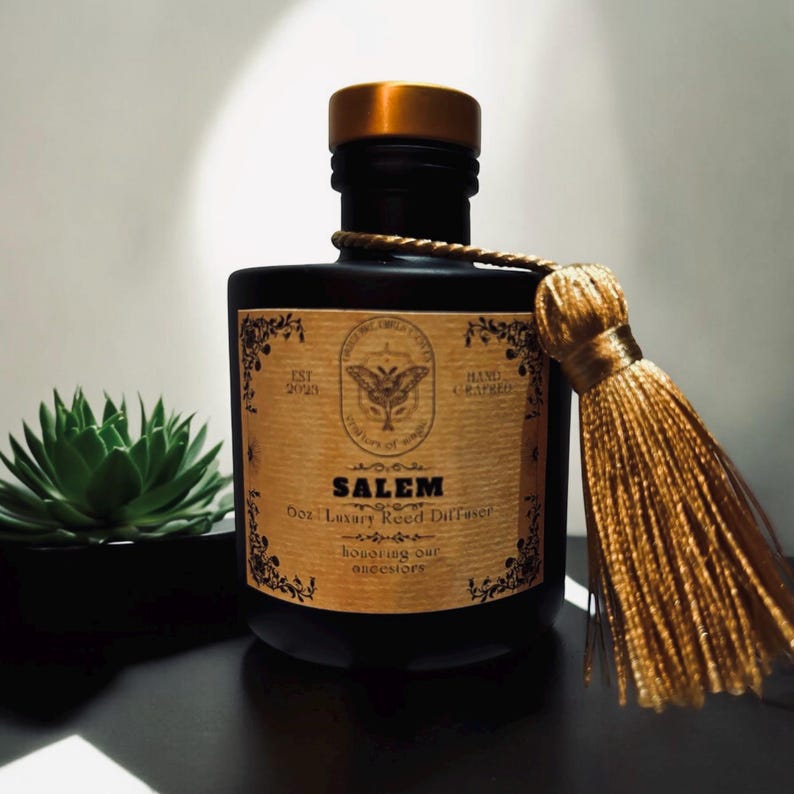 May include: A black glass bottle with a gold lid and a gold tassel. The bottle has a label that says "Salem Dot Luxury Reed Diffuser Honoring our ancestors" with a butterfly graphic.