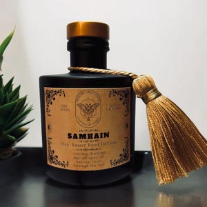May include: A black glass bottle with a gold lid and a gold tassel. The bottle has a label that says "Samhain 6oz Luxury Reed Diffuser Casting shadows that whisper of ancient tales through the veil".