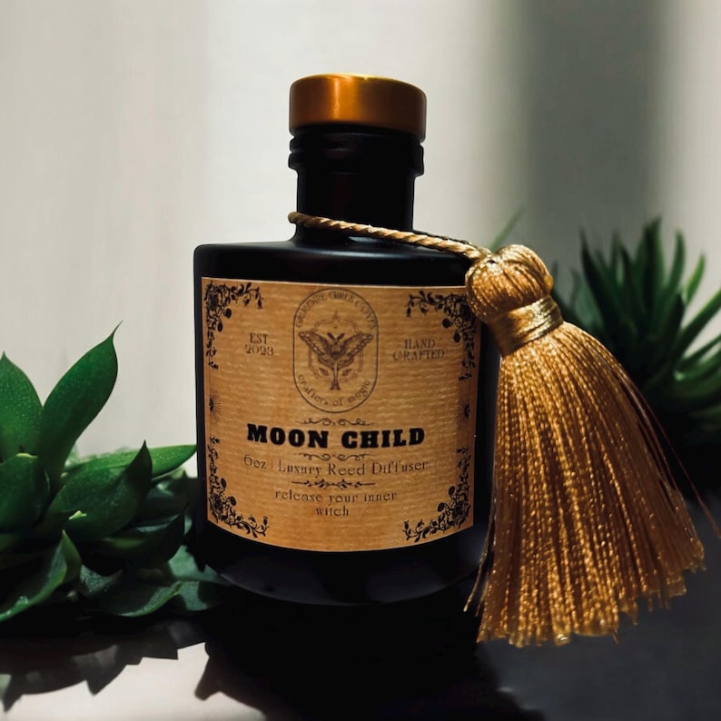 May include: A black glass bottle with a gold lid and a gold tassel. The bottle has a label that says "Moon Child 6oz Luxury Reed Diffuser release your inner witch".