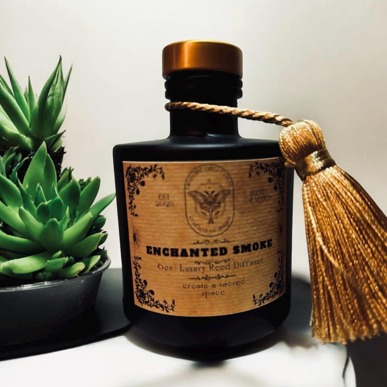 May include: A black glass bottle with a gold lid and a gold tassel. The bottle has a label that says "Enchanted Smoke 6oz Luxury Reed Diffuser create a sacred space".