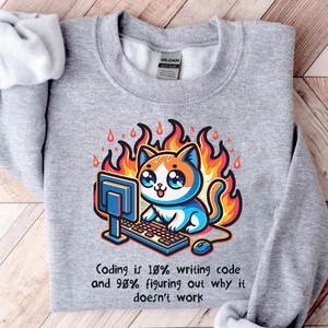 May include: Gray sweatshirt featuring a cartoon cat with orange and blue fur, working on a computer with flames around it. The text reads, "Coding is 10% writing code and 90% figuring out why it doesn't work."
