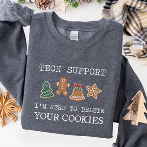 May include: Dark heather gray sweatshirt with the text "TECH SUPPORT" and "I'M HERE TO DELETE YOUR COOKIES". The design includes gingerbread men, a Christmas tree, a bell, and a star. A wooden tree is on the sleeve.