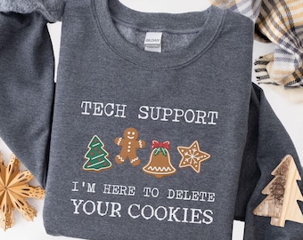 May include: Dark heather gray sweatshirt with the text "TECH SUPPORT" and "I'M HERE TO DELETE YOUR COOKIES". The design includes gingerbread men, a Christmas tree, a bell, and a star. A wooden tree is on the sleeve.