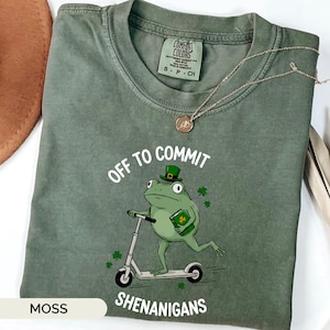 May include: A moss green t-shirt with the words "OFF TO COMMIT SHENANIGANS" and a graphic of a frog riding a scooter and wearing a leprechaun hat. A gold necklace is draped across the shirt.
