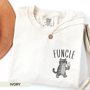 May include: An ivory t-shirt featuring a graphic of a grey tabby cat wearing black sunglasses and holding an ice cream cone. Above the cat, the word "FUNCLE" is printed in black. The shirt has a Comfort Colors brand label at the neck.
