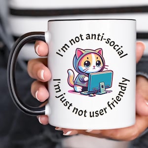 May include: White ceramic mug with a black interior and handle. The mug features a cartoon cat wearing a hoodie and working on a computer. Text on the mug reads "I'm not anti-social, I'm just not user friendly."