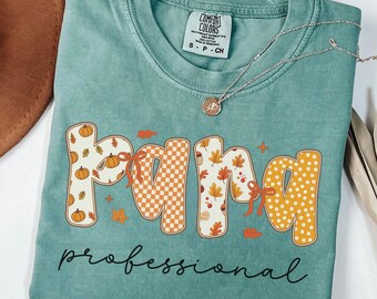 May include: Light green t-shirt with the word "PAMA" in a fall-themed design, featuring pumpkins, leaves, and orange bows. The word "professional" is written below in black script. A silver necklace with a pendant is draped across the shirt.
