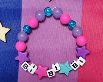 May include: A pink, purple, and blue beaded bracelet with the word "B I B I" spelled out in colorful letter beads. The bracelet is on a background of a pink, blue, and purple fabric.