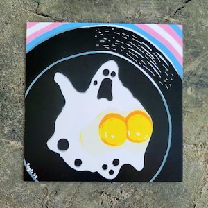 May include: A white ghost-shaped fried egg with two yellow yolks in a black pan. The pan has a white rim and is set against a pink, blue, and white striped background.