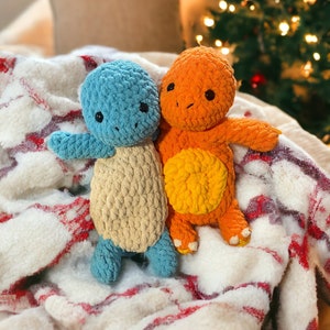 May include: Two crocheted stuffed toys, one blue and one orange, are lying on a white and red blanket. The toys are shaped like turtles and have embroidered eyes.