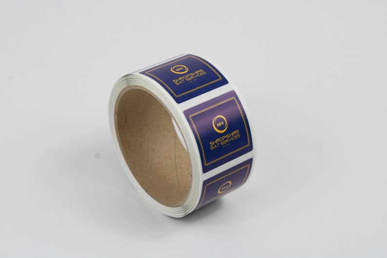 May include: A roll of square stickers with a navy blue background and gold trim. Each sticker features the text "SHROPSHIRE BAY SERVICES" and a gold circular logo. The stickers are on a white background.