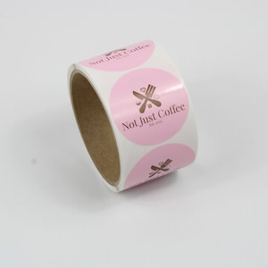 May include: A roll of round stickers with a pink background and the text "Not Just Coffee" in brown. The stickers also feature a brown graphic of a fork and knife. The roll is on a white surface.