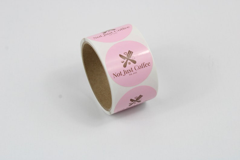 May include: A roll of round stickers with a pink background and the text "Not Just Coffee" in brown. The stickers also feature a brown graphic of a fork and knife. The roll is on a white surface.