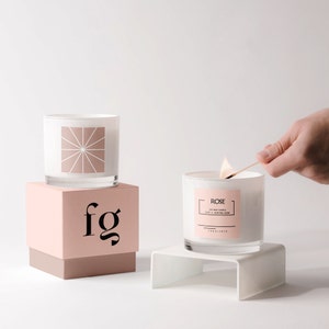 May include: Two white glass candles, one on a pink box with "fg" printed on it, and the other on a white stand. The candle on the right is being lit with a match. The candle on the right has a label that says "Rose".