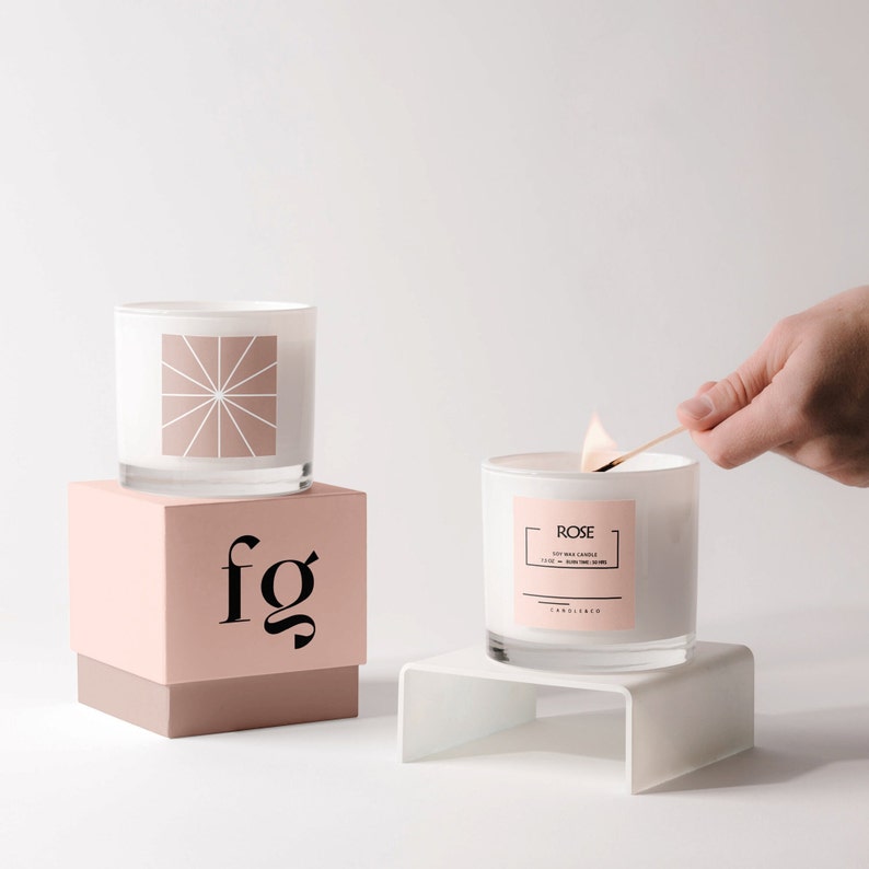 May include: Two white glass candles, one on a pink box with "fg" printed on it, and the other on a white stand. The candle on the right is being lit with a match. The candle on the right has a label that says "Rose".