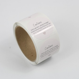May include: A roll of white, round stickers with the text "Contains" and a list of potential allergens in a script font. The stickers have a light pink heart and are for food products.