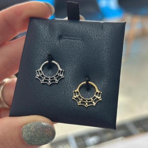 May include: Two septum rings on a black display. One is silver, the other gold. Each ring features a circular design with a decorative web pattern. The rings are designed for nose piercings.