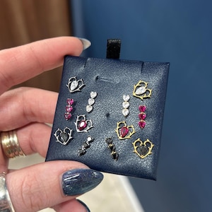 May include: A selection of stud earrings presented on a navy blue card. The earrings showcase heart and teardrop shapes, adorned with clear, pink, and black gemstones. Some have gold or silver settings.