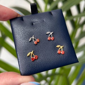 May include: Four pairs of cherry stud earrings. Each earring features a pair of red gemstone cherries with a silver or gold-coloured stem and leaf design. The earrings are displayed on a dark blue jewellery card.
