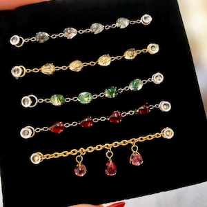 May include: Five gold-toned metal jewellery connectors featuring various gemstone designs. Four connectors feature a linear arrangement of teardrop or oval stones in clear, golden, green, and red hues. The fifth connector features three dangling pink stones.
