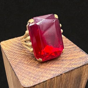 May include: A gold-toned ring featuring a large, rectangular, faceted, red gemstone. The ring is set on a wooden block against a black background. The gemstone is the focal point, catching the light and showcasing its vibrant color.
