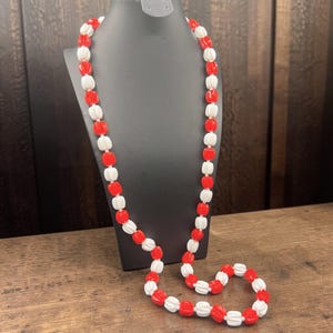 May include: A long necklace with alternating red and white beads. The beads are cylindrical with a ribbed texture. The necklace is displayed on a black mannequin against a wooden background.