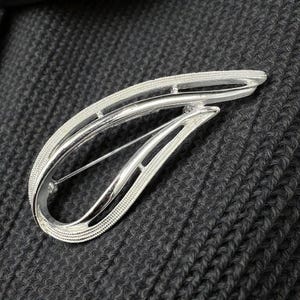 May include: A silver brooch with a flowing, abstract shape. The brooch has a pin fastening on the reverse. The design incorporates a textured edge and a smooth, open central section, offering a contemporary and stylish accessory.