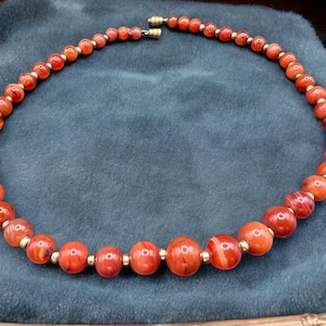 May include: A necklace made of carnelian beads with gold accents. The beads are a deep orange color with white and red streaks.
