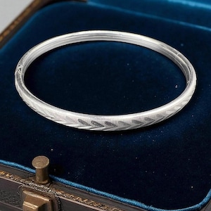 Peut inclure: Un bracelet jonc argenté avec un motif géométrique. Le bracelet est présenté à l'intérieur d'un écrin à bijoux vintage avec un intérieur en velours bleu. Le jonc présente un motif de chevrons répétés.