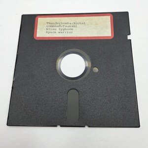 Puede incluir: Un disquete negro de 3,5 pulgadas con una etiqueta que presenta el texto "Thunderbombs: Rocket, command: Tsunami, Alien typhoon, Space warrior". El disco tiene una abertura circular central y una ranura rectangular.