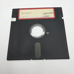 Puede incluir: Un disquete negro de 3,5 pulgadas con un cubo plateado y una ranura rectangular. Una etiqueta con las palabras "Threshold" y "sil" está adherida en la parte superior. La etiqueta está bordeada por un rectángulo rojo.