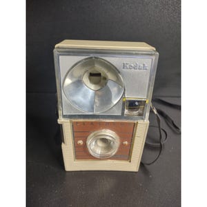 Könnte beinhalten: Vintage Kodak Flashmite Kamera mit cremefarbenem und braunem Gehäuse. Die Kamera hat einen silbernen Blitzreflektor und eine klare Linse. Das Wort "Kodak" ist oben aufgedruckt, "Flashmite" auf der Vorderseite. Ein schwarzer Riemen ist befestigt.