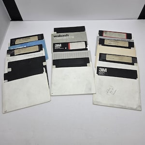 May include: A collection of vintage 5.25-inch floppy disks, featuring black and beige disks with labels. The disks are in white paper sleeves, some with printed text. Brands include 3M and Wabash.
