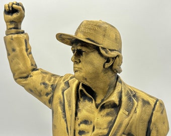 May include: A gold-colored bust sculpture of a person wearing a black suit jacket and a baseball cap with the text "Make America Great Again". The person's right arm is raised in a fist.