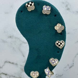 Puede incluir: Un conjunto de seis pendientes de tono dorado con un patrón a cuadros en varios colores, incluyendo negro, blanco, rosa y morado. Los pendientes se muestran en un expositor de terciopelo verde.