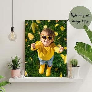 May include: A child wearing yellow heart-shaped sunglasses and a yellow jumper stands in a field of green grass with yellow leaves scattered around them.