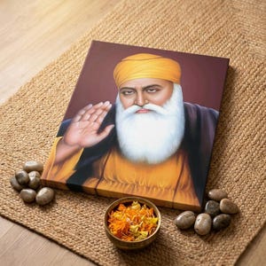 Könnte beinhalten: Leinwanddruck mit einem Porträt einer religiösen Figur mit weißem Bart und gelbem Turban. Das Bild ist vor einem weinroten Hintergrund platziert, mit dekorativen Steinen und einer kleinen Schale mit Blütenblättern auf einer gewebten Matte.