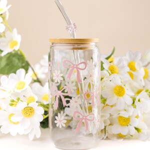 May include: A clear glass tumbler with a bamboo lid and a glass straw. The tumbler is decorated with pink bows and white daisy flowers. The background features white daisies with yellow centers.