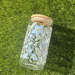 May include: A clear glass tumbler with a bamboo lid featuring a printed pattern of light blue flowers, small yellow centers, and pale green leaves. The cylindrical glass container has a smooth surface and a flat base, designed for holding beverages.