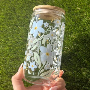 May include: A clear glass tumbler featuring a floral pattern of light blue and white flowers with yellow centers and pale green leaves. The cylindrical glass container is topped with a circular bamboo lid that fits securely over the opening.