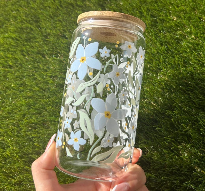 May include: A clear glass tumbler featuring a floral pattern of light blue and white flowers with yellow centers and pale green leaves. The cylindrical glass container is topped with a circular bamboo lid that fits securely over the opening.
