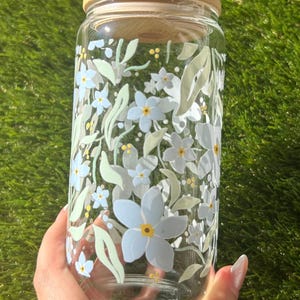 May include: A clear glass tumbler with a bamboo lid featuring a printed pattern of light blue flowers, pale green leaves, and small yellow dots. The cylindrical glass container has a smooth surface and a wide opening, showcasing a botanical design.