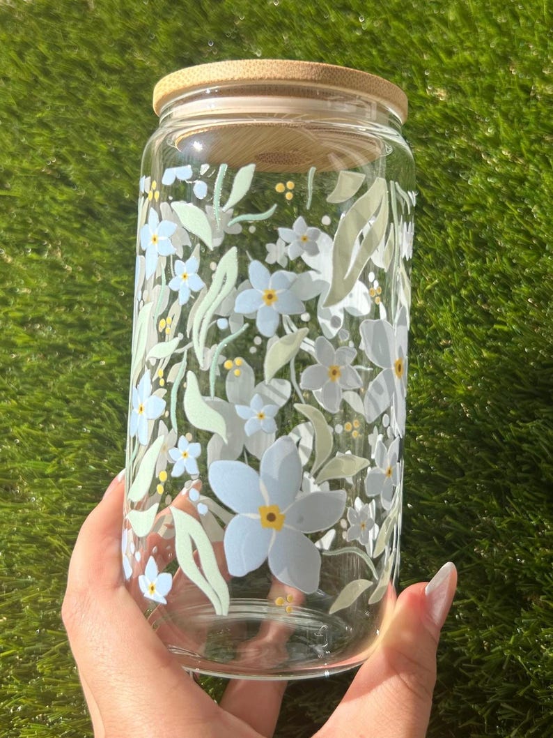 May include: A clear glass tumbler with a bamboo lid featuring a printed pattern of light blue flowers, pale green leaves, and small yellow dots. The cylindrical glass container has a smooth surface and a wide opening, showcasing a botanical design.