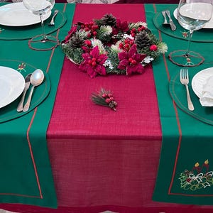 May include: A festive Christmas table setting with a green and red color scheme. The table is set with white plates, silverware, and wine glasses. A decorative wreath and embroidered details add to the holiday theme.