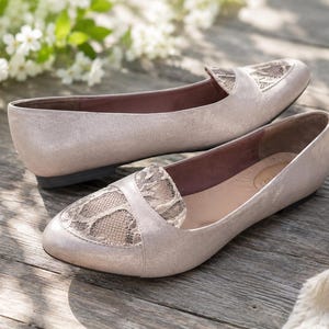May include: A pair of light beige metallic flats featuring a rounded toe and a decorative lace overlay panel across the vamp. The shoes have a low profile, a smooth finish, and a solid sole, showcasing a neutral color palette and a slip-on design.
