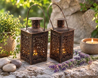 Puede incluir: Dos faroles cuadrados de metal con acabado en bronce oscuro y patrones intrincados recortados en los cuatro lados. Cada farol cuenta con un asa superior, una chimenea cilíndrica con perforaciones circulares y una vela encendida en su interior.
